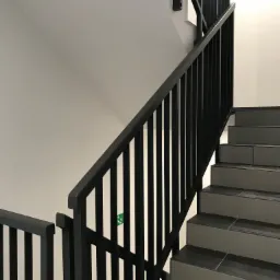 Schody z czarną metalową balustradą i szarymi płytkami, widoczny znak ewakuacyjny.