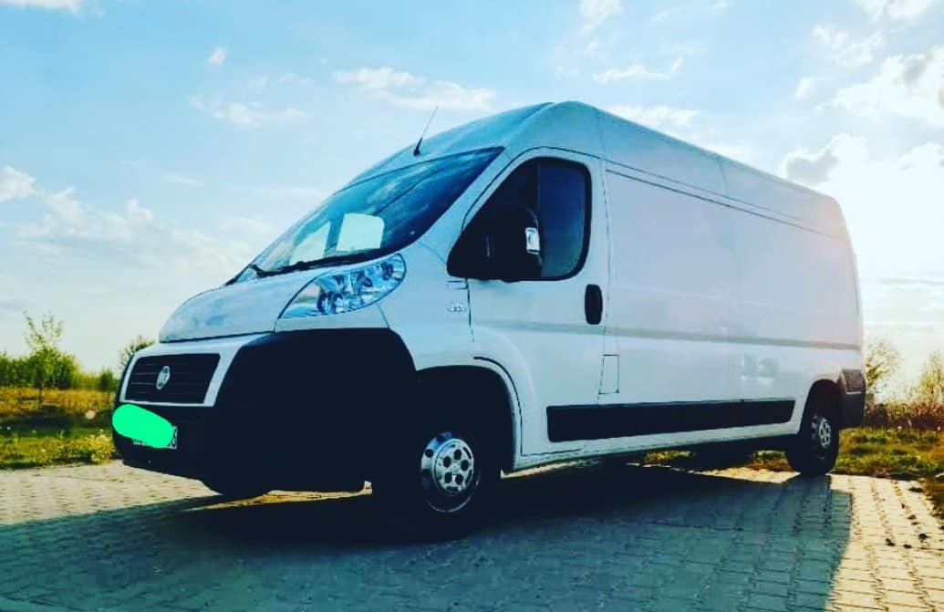 Biały samochód dostawczy Fiat Ducato na brukowanej powierzchni w Warszawie, z błękitnym niebem w tle. Widoczne chromowane kołpaki i delikatne odbicie światła na karoserii.