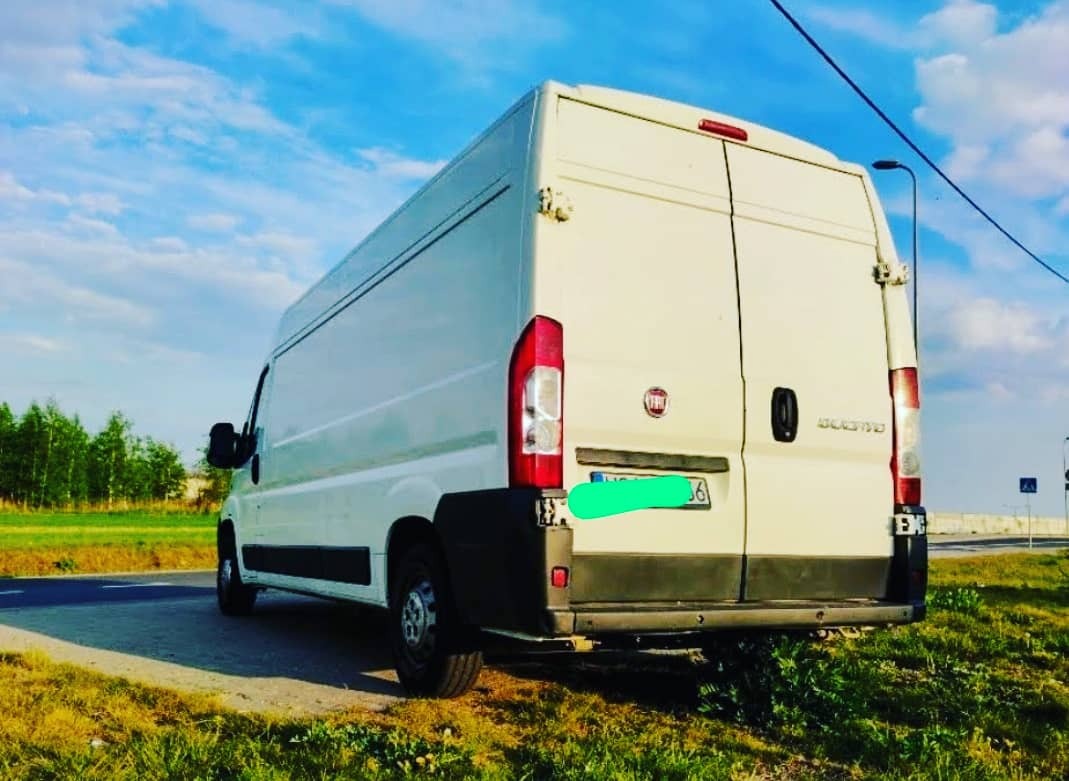 Biały samochód dostawczy Fiat Ducato zaparkowany na poboczu drogi z widocznymi tablicami rejestracyjnymi, zielonym trawnikiem i błękitnym niebem w tle.
