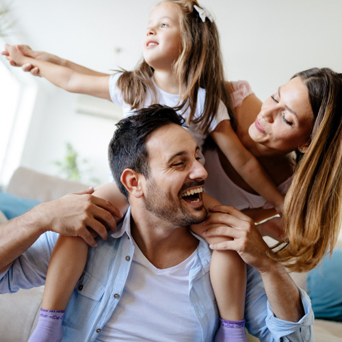 Uśmiechnięty ojciec z córką na barana, obok matka w jasnym wnętrzu mieszkalnym.