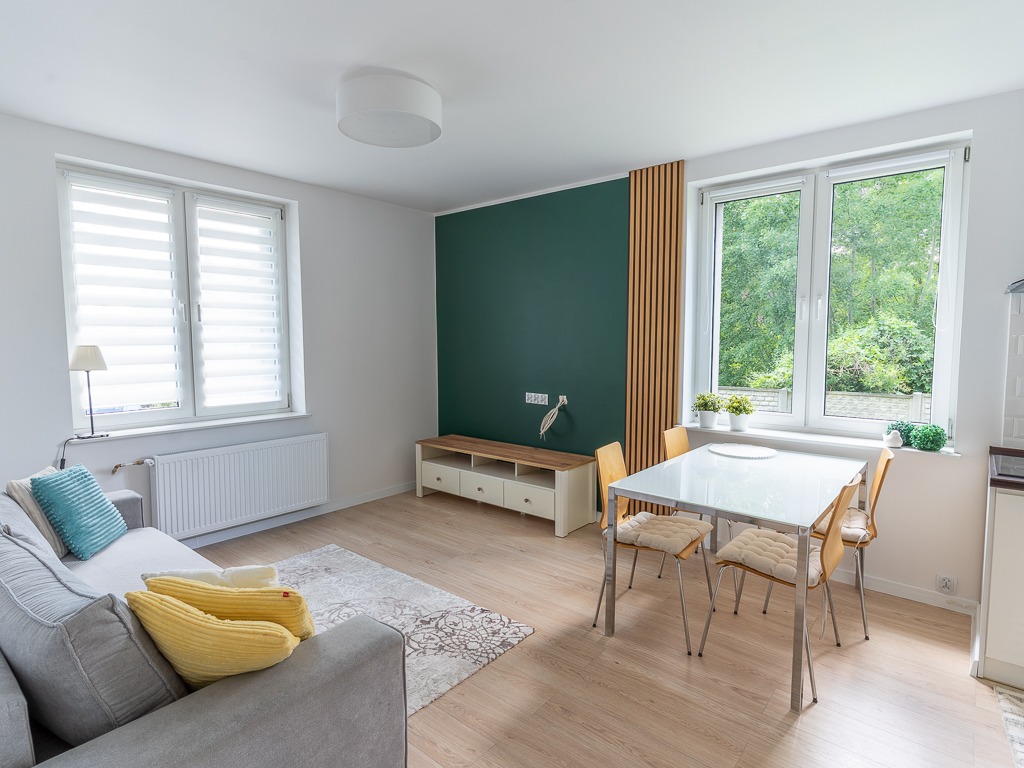 Jasne wnętrze mieszkania z szarą sofą, białym stołem i zieloną ścianą. Widok na okno z roletami i drewniane panele dekoracyjne. Minimalistyczny design.