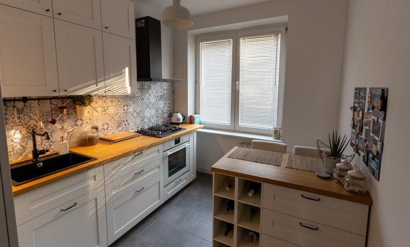 Jasna kuchnia z drewnianym blatem i wzorzystą mozaiką nad blatem. Widoczne okno z żaluzjami i stół z miejscami do siedzenia. W tle zdjęcia pamiątki na ścianie.