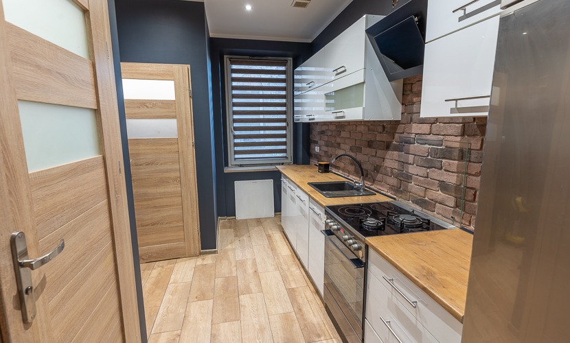 Nowoczesna kuchnia z białymi szafkami, drewnianymi blatami i ceglaną ścianą. Widoczny fragment lodówki, okno z żaluzjami i drewniane drzwi z przeszkleniami.