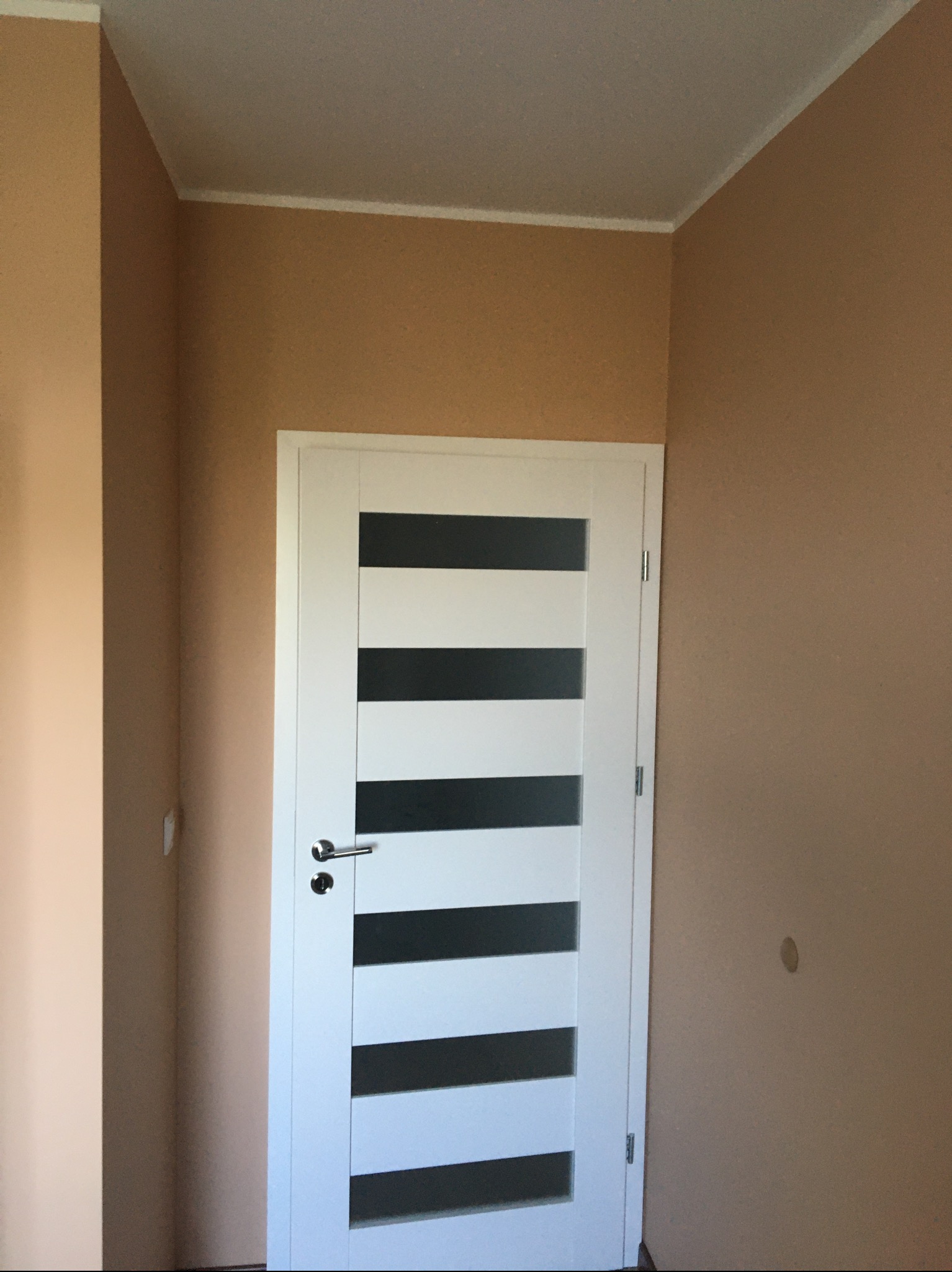 Ściana pomalowana na beżowo, z białą, nowoczesną stolarką drzwiową z przeszkleniami w postaci poziomych pasów. Widoczny fragment sufitu z listwą wykończeniową. Na ścianie widoczne ślady po...