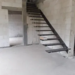 Surowe metalowe schody z prostymi stopniami, zamontowane w surowym wnętrzu budynku z białych bloczków, widok z dołu na konstrukcję w trakcie montażu.