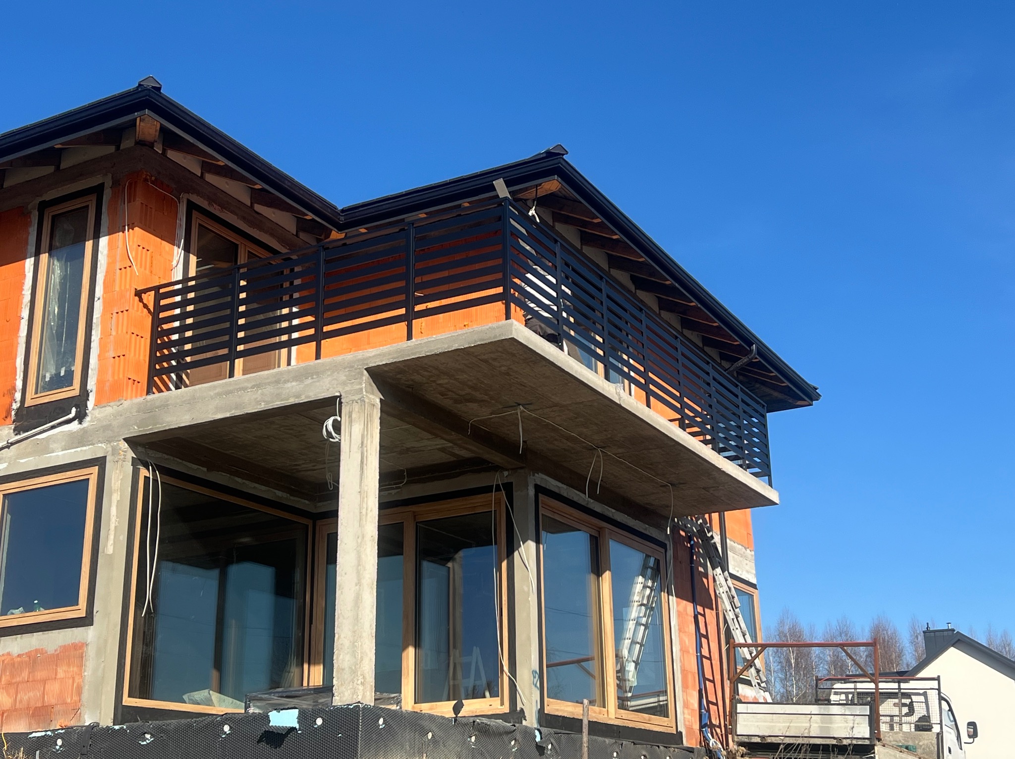 Surowy stan budynku w trakcie budowy z widocznymi oknami z drewnianą ramą, betonowym balkonem z czarną metalową balustradą i fragmentem drabiny opartą o ścianę.
