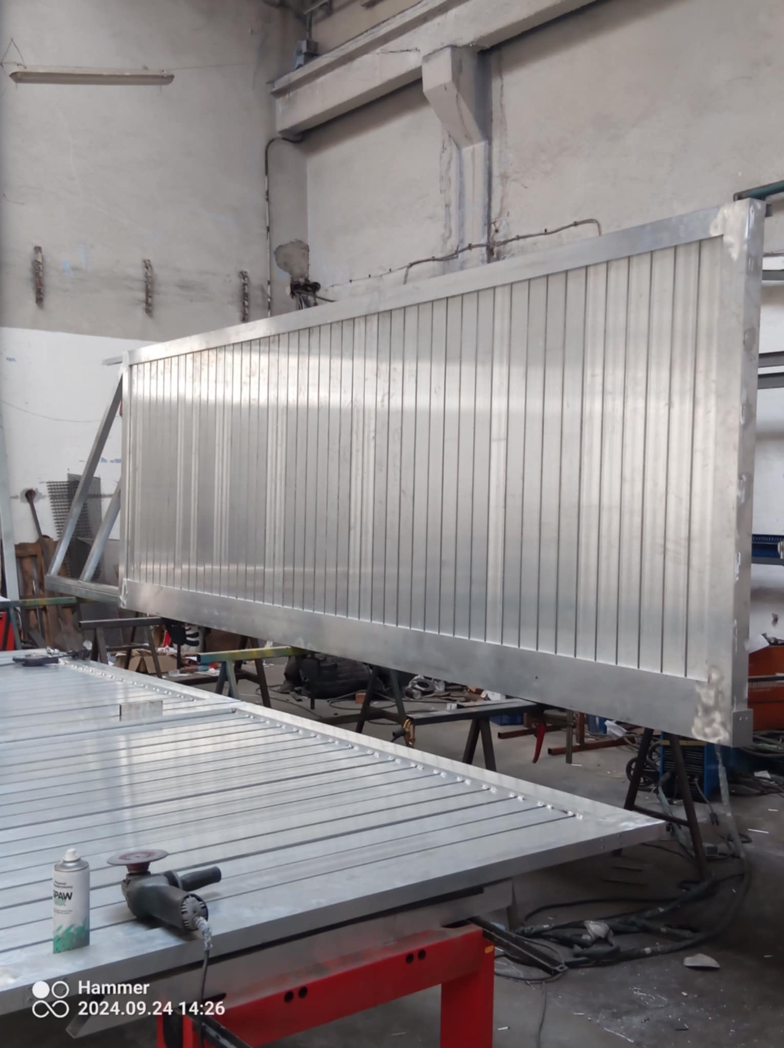 Wnętrze warsztatu z częściowo ukończoną, dużą, metalową bramą przesuwną z pionowymi panelami, leżącą na stole roboczym obok szlifierki kątowej i aerozolu, widoczne ślady spawania.