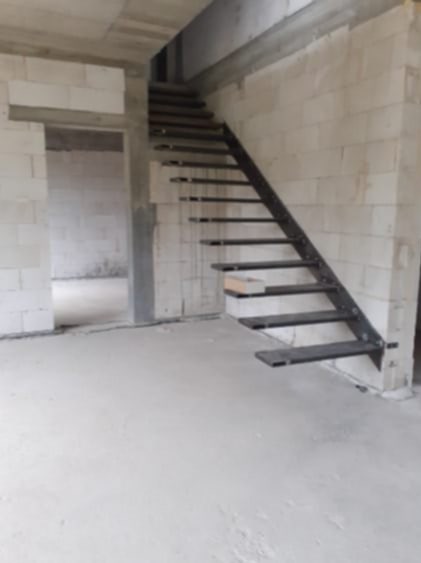 Surowe metalowe schody z prostymi stopniami, zamontowane w surowym wnętrzu budynku z białych bloczków, widok z dołu na konstrukcję w trakcie montażu.