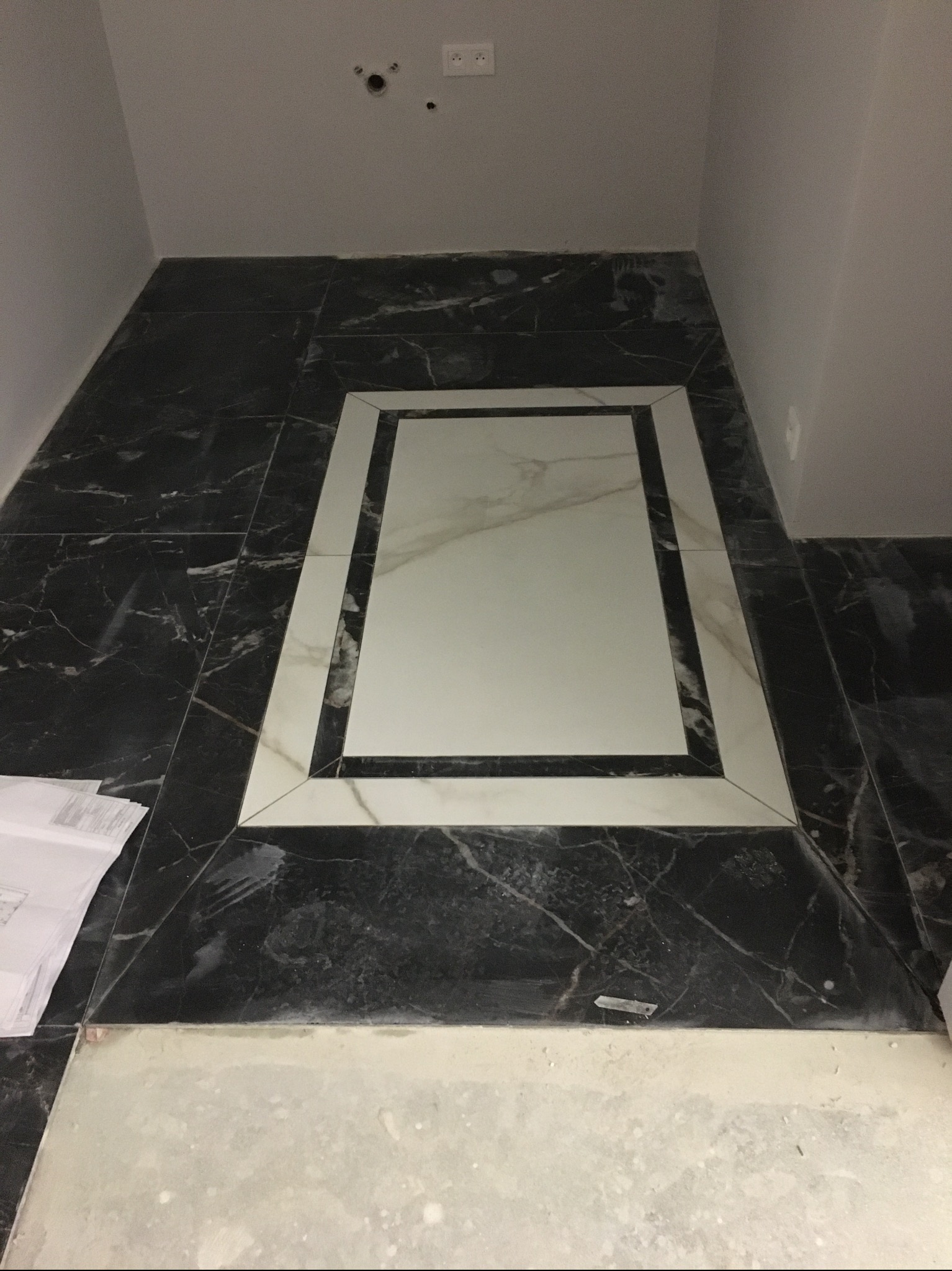 Ułożona posadzka z czarnych i białych płytek marmurowych z dekoracyjnym prostokątnym wzorem na środku, widoczne plany na podłodze i gniazdka elektryczne na ścianie.