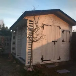 Biała drewniana altana z czarnymi okuciami i okiennicami, oświetlona popołudniowym słońcem, z młodym drzewkiem na pierwszym planie i betonowym płotem w tle.