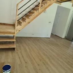 Drewniane schody z czarnymi metalowymi barierkami w jasnym wnętrzu domu, widoczna puszka z farbą na podłodze.