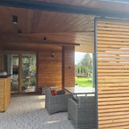 Zadaszony taras z drewnianą elewacją, meblami ogrodowymi z rattanu i drewnianą ścianką działową z poziomych listew, widok na ogród z huśtawką.