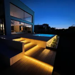 Oświetlony taras z drewnianą podłogą i schodami prowadzącymi do małego basenu przy nowoczesnym domu w Podolszynie Nowym, sfotografowany o zmierzchu.