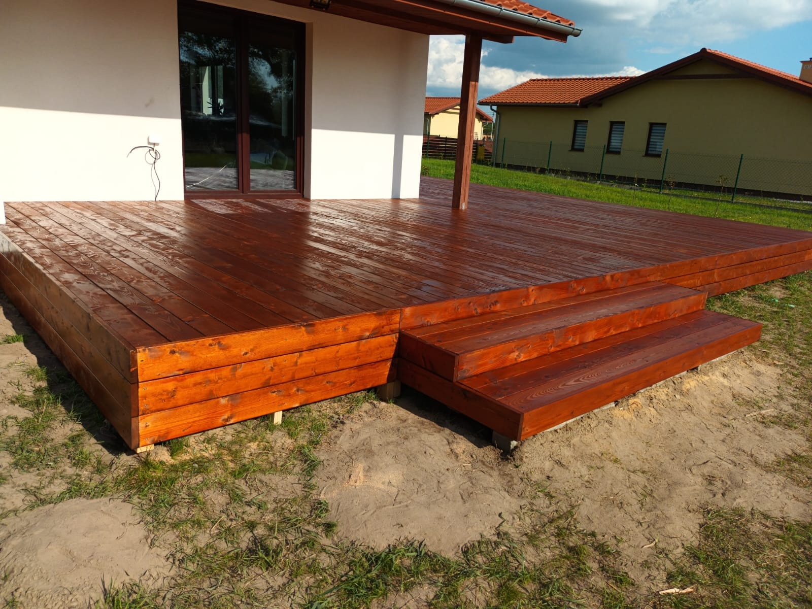 Nowoczesny taras z drewna, świeżo polakierowany na ciemny brąz, z niskimi schodkami prowadzącymi na podest, na tle białego domu z ciemnymi oknami i zielonego trawnika.