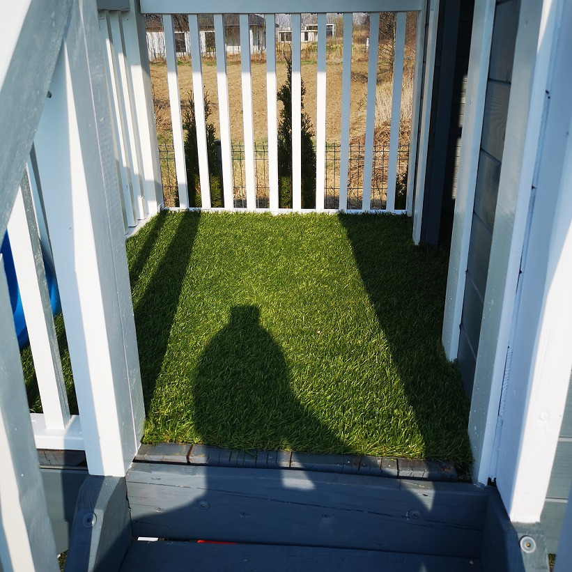 Widok z góry na fragment konstrukcji z białymi balustradami i sztuczną trawą wewnątrz, cień osoby fotografującej na trawie.