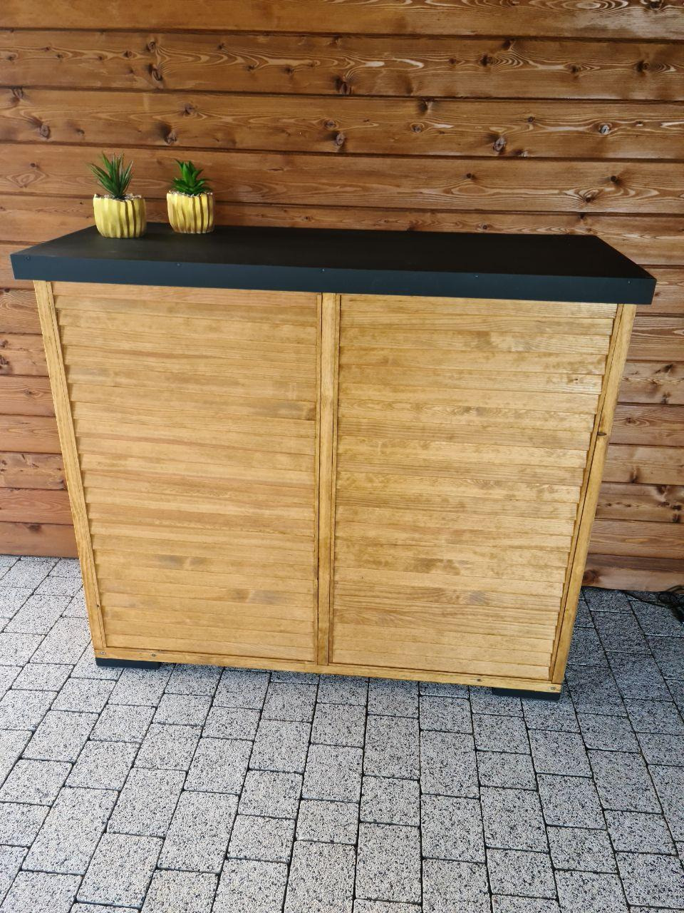 Drewniana szafka ogrodowa z lamelowymi drzwiczkami, czarnym blatem i dwoma złotymi doniczkami z sukulentami na tle drewnianej ściany i szarej kostki brukowej.