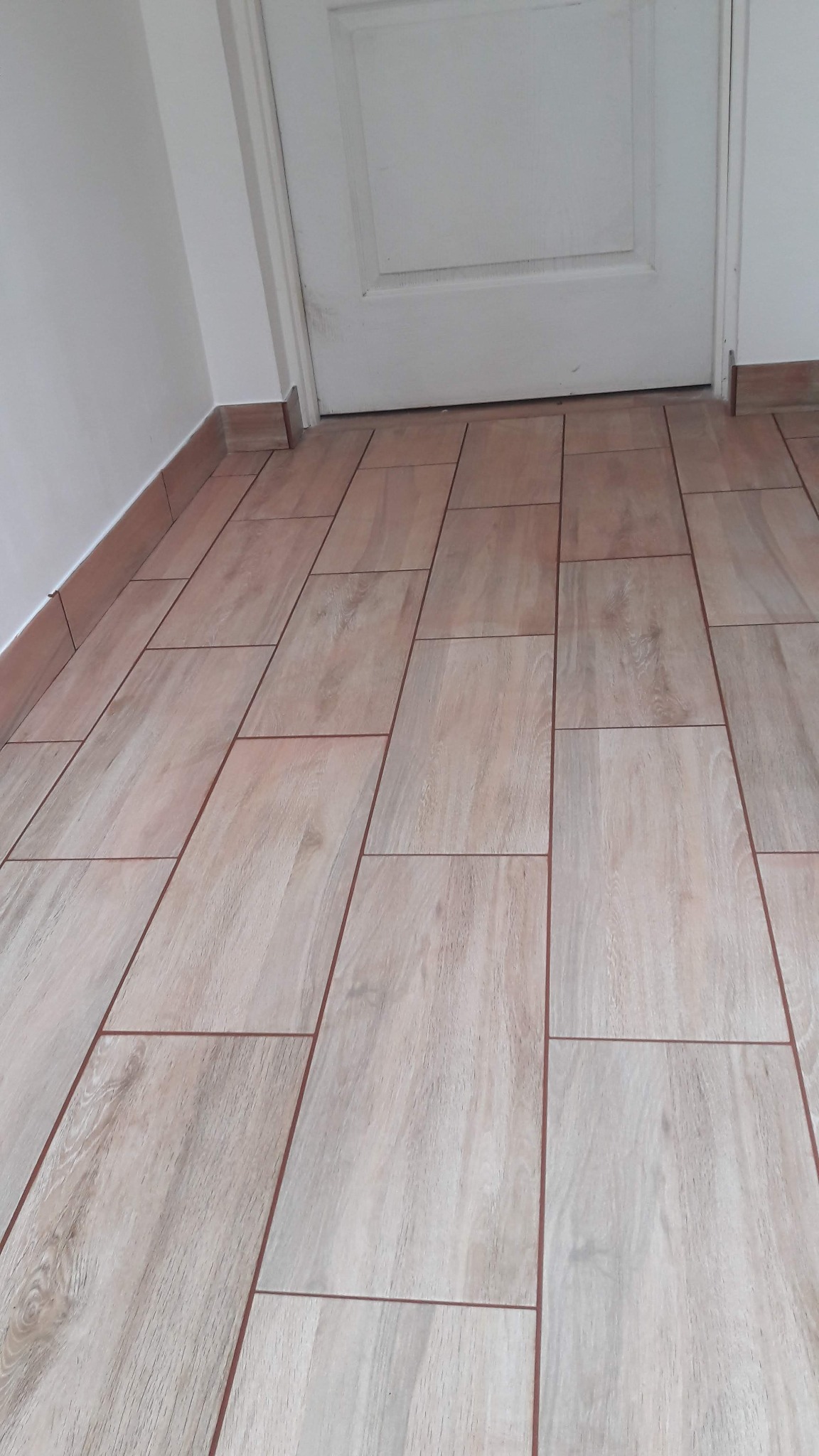 Ułożone podłużne płytki podłogowe imitujące drewno, z widoczną fugą w kolorze brązowym, dopasowane do listwy przypodłogowej przy białej ścianie i białych drzwiach.