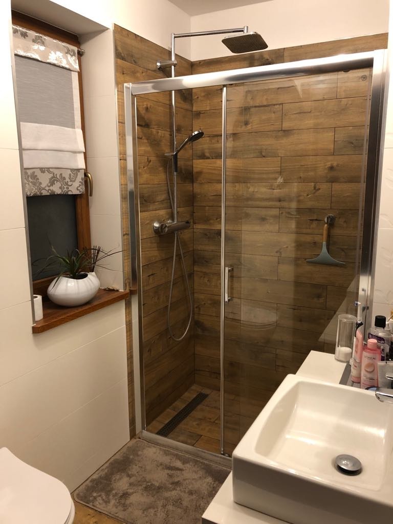 Nowoczesna łazienka z kabiną prysznicową wykończoną płytkami imitującymi drewno, widoczny fragment umywalki i okno z roletą rzymską.