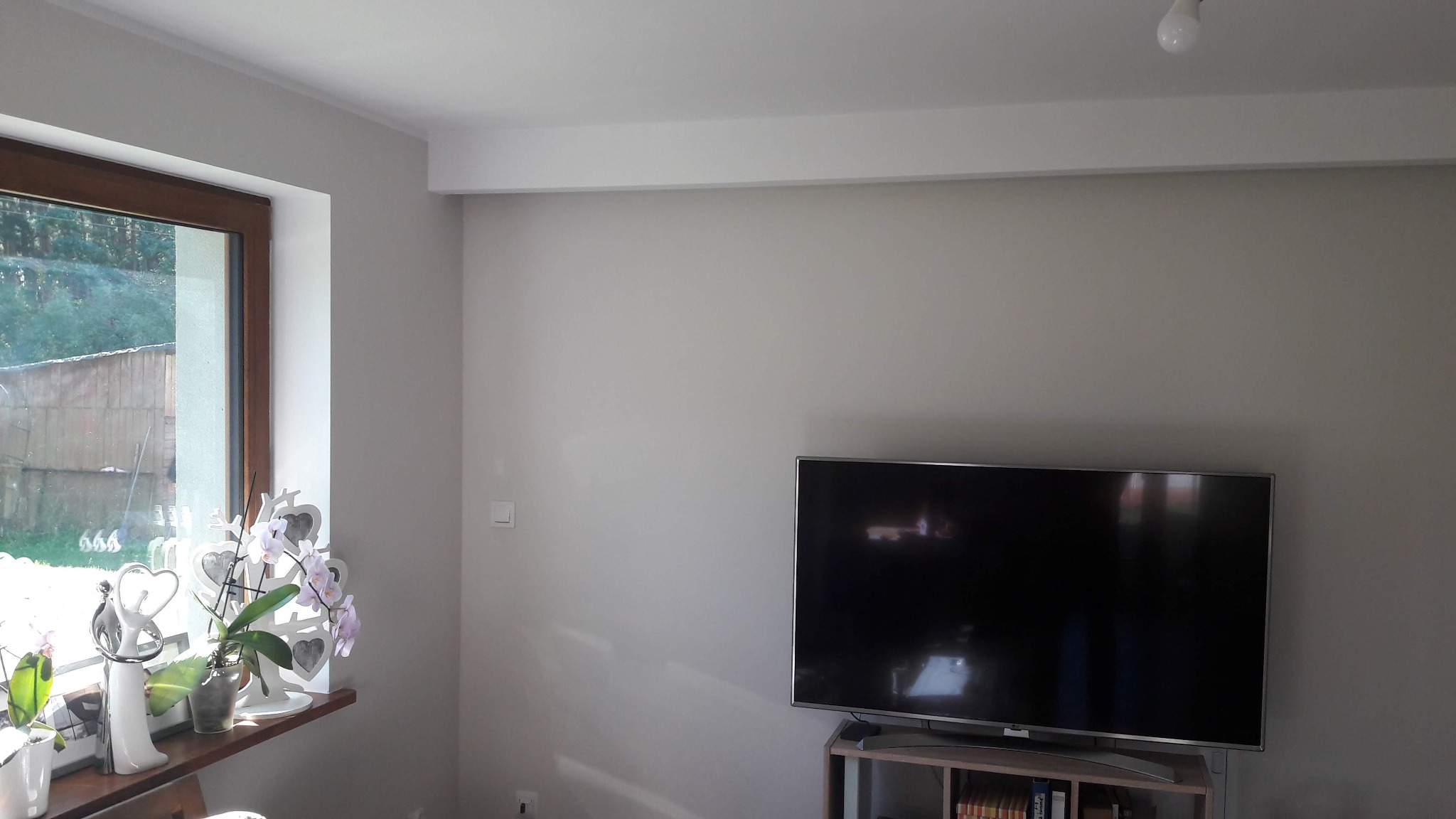 Jasne wnętrze pokoju po remoncie, ściany pomalowane na szaro, widoczne okno z drewnianą ramą, parapet z dekoracjami i kwiatami, oraz telewizor na szafce. Listwa sufitowa i gładkie ściany.