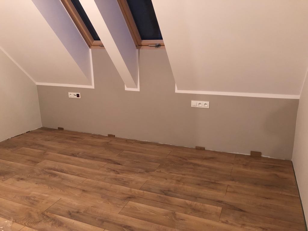 Układanie paneli podłogowych w pokoju na poddaszu, widoczne kliny dystansowe przy ścianie, ściana pomalowana na dwa kolory, okno dachowe.
