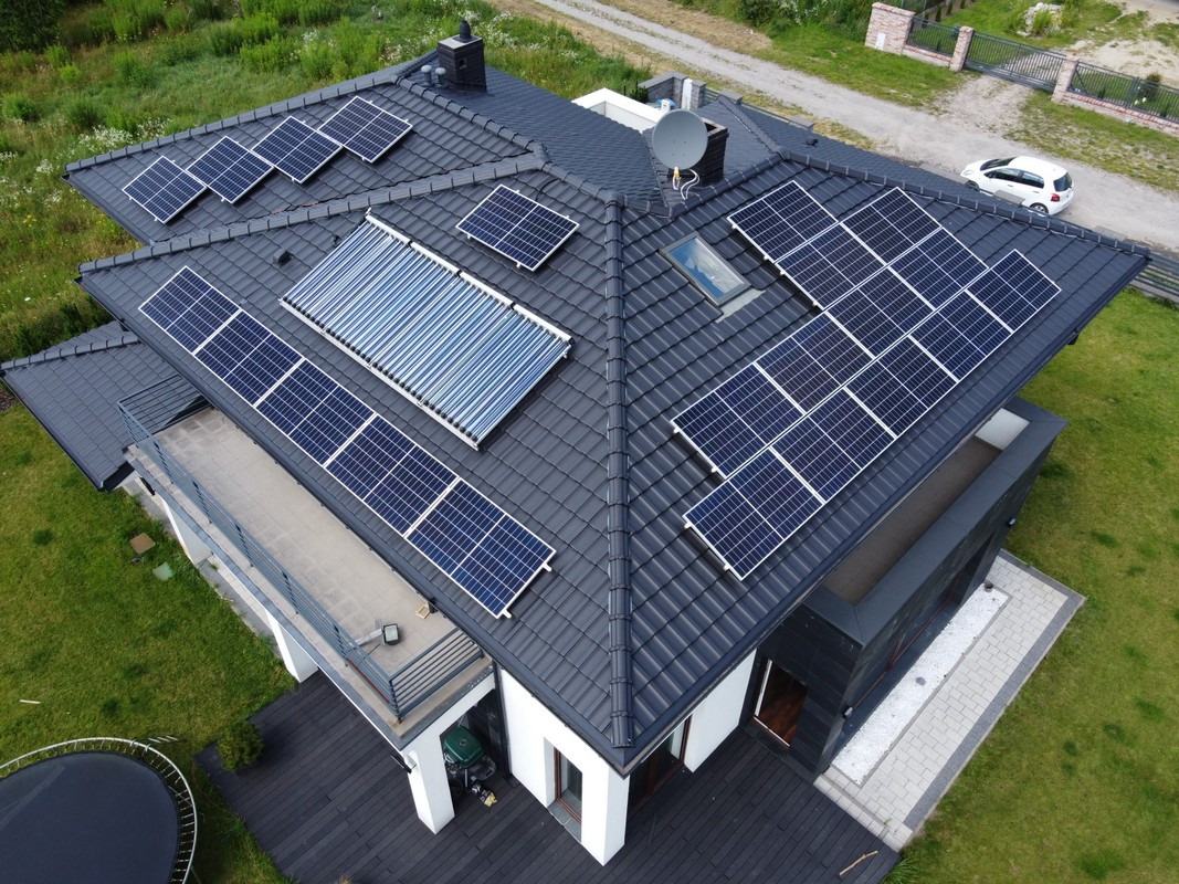 Widok z góry na dach domu z zainstalowanymi panelami słonecznymi i kolektorami słonecznymi, antena satelitarna, okno dachowe, na podwórku trampolina i zaparkowany samochód.