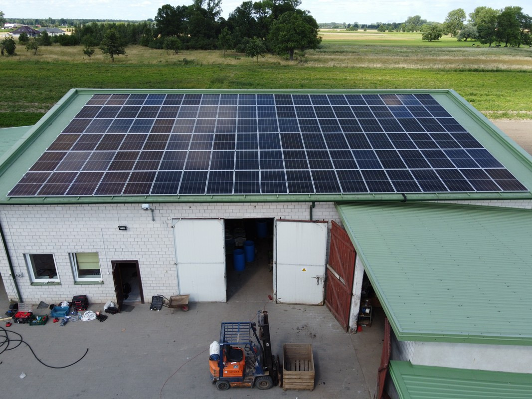 Widok z góry na budynek z panelami słonecznymi na dachu, z otwartymi drzwiami garażowymi, w tle zielone pole i las, w pobliżu drzwi widać wózek widłowy i narzędzia.
