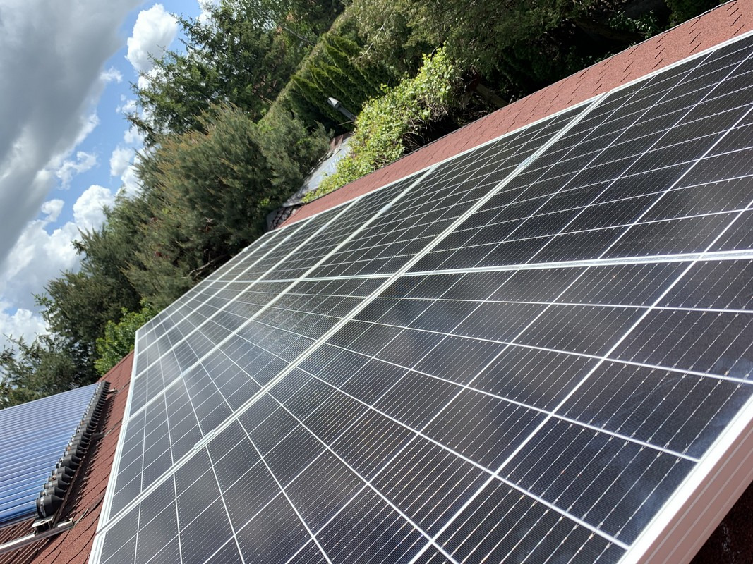 Panele słoneczne na brązowym dachu domu, częściowo zacienione przez chmury i otoczone zielenią drzew.