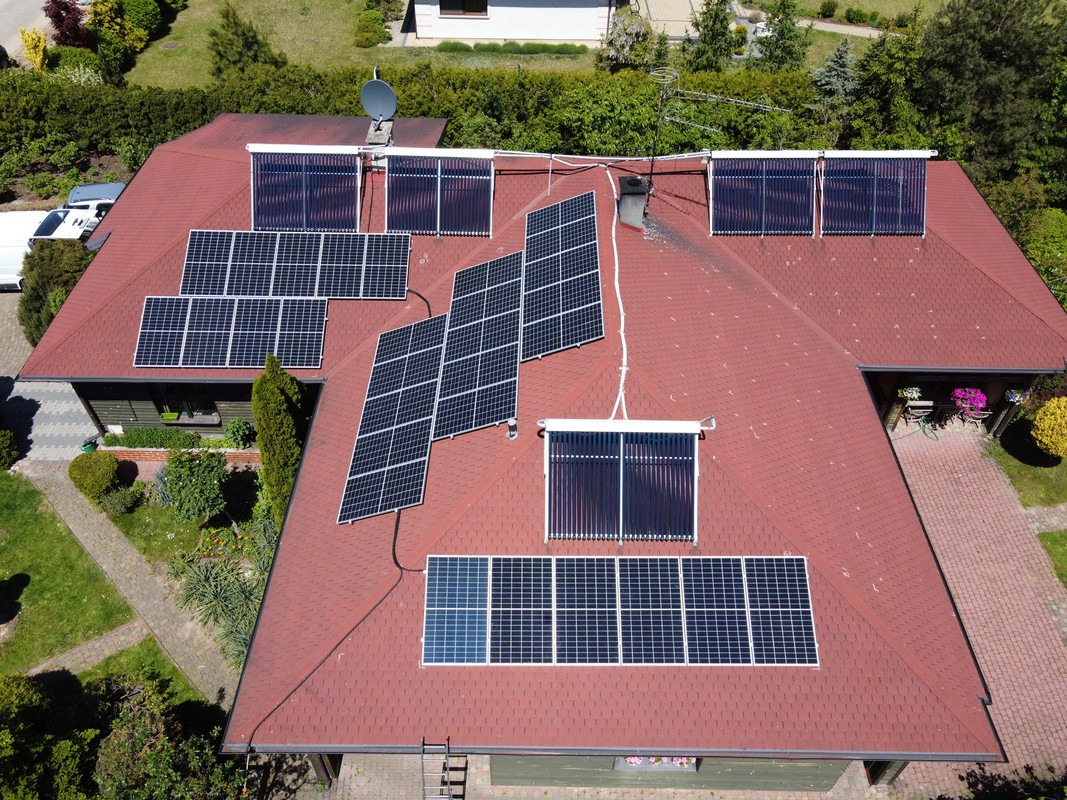 Widok z lotu ptaka na dom z czerwonym dachem pokryty panelami fotowoltaicznymi i kolektorami słonecznymi; widoczna antena satelitarna, drabina oparta o ścianę i zadbany ogród.
