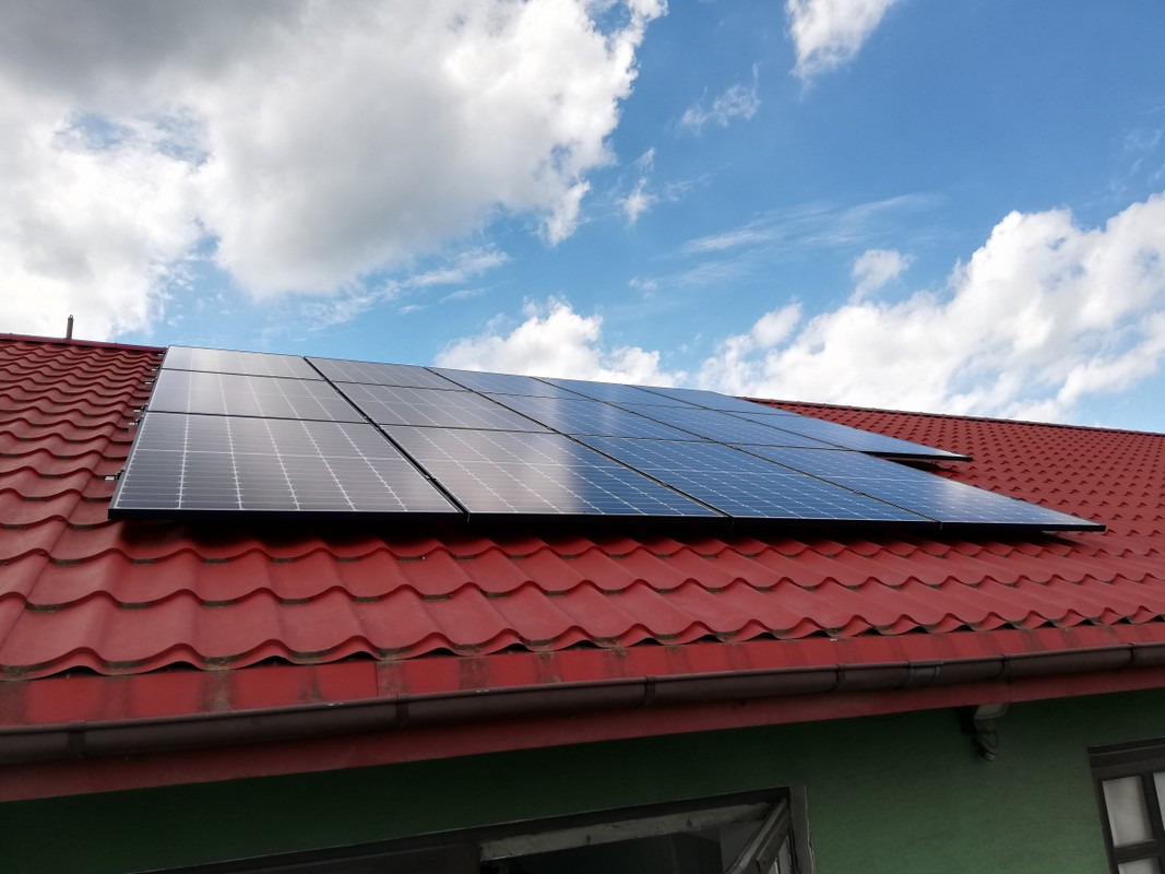 Panel fotowoltaiczny na dachu z czerwonej dachówki, widok z dołu na tle błękitnego nieba z chmurami.
