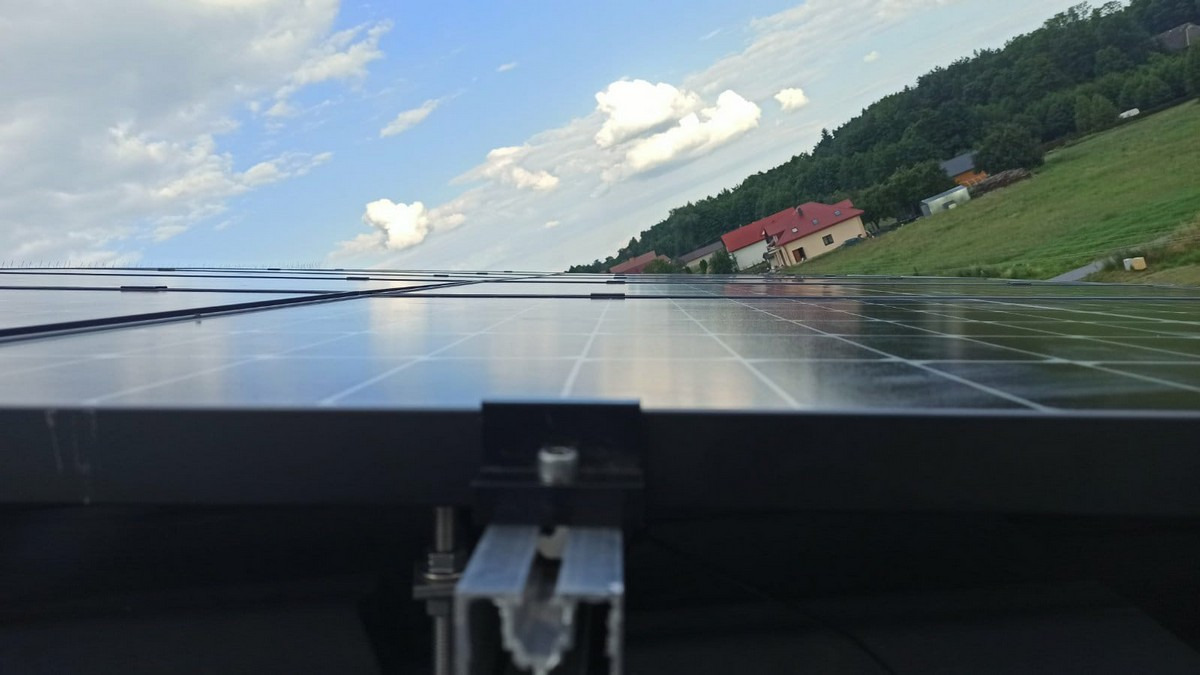 Panele słoneczne w Poznaniu zamontowane na dachu budynku, widoczne mocowania i fragment krajobrazu z zielonym wzgórzem i domami w tle.