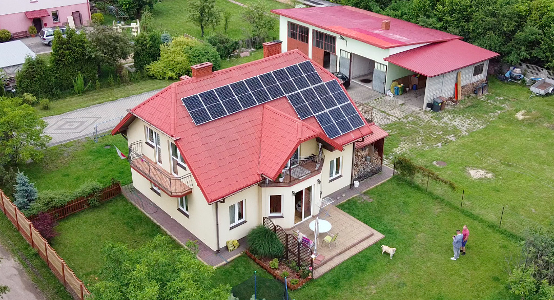 Dom jednorodzinny z czerwonym dachem i panelami słonecznymi, widok z góry, ogród z trawnikiem, spacerujący ludzie i pies.