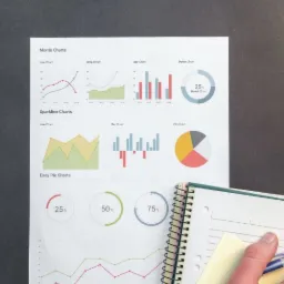 Analiza danych finansowych: Wykresy i notatnik na biurku z laptopem, ręka z długopisem zaznacza informacje.