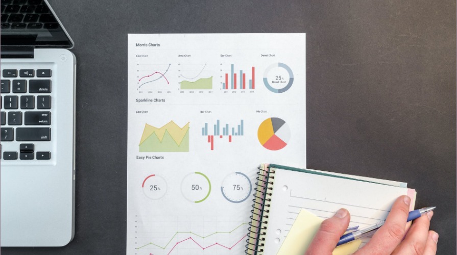 Analiza danych finansowych: Wykresy i notatnik na biurku z laptopem, ręka z długopisem zaznacza informacje.