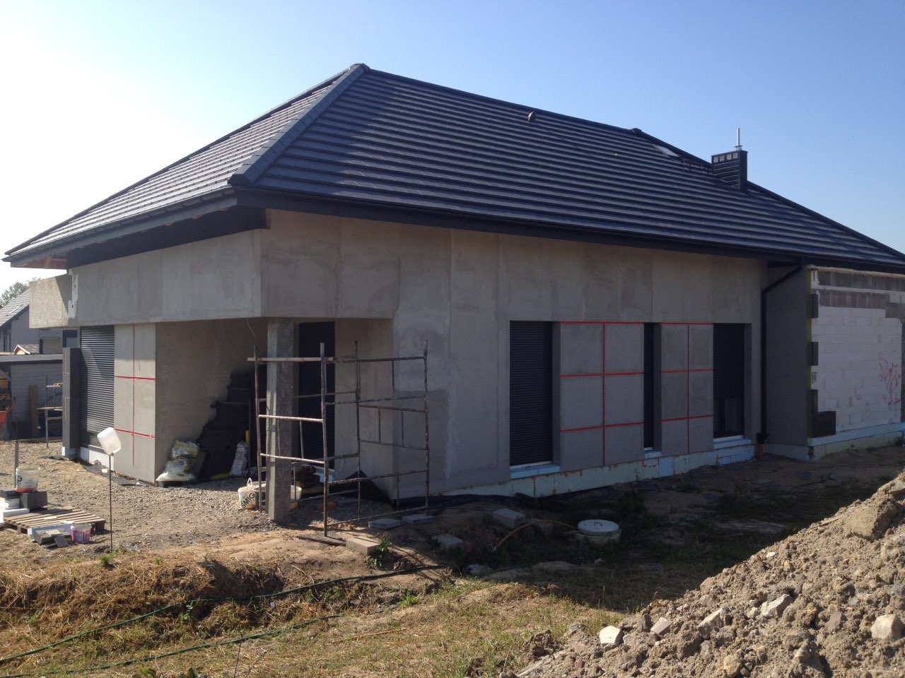 Dom w trakcie budowy: surowe ściany zewnętrzne z widocznymi elementami izolacji, czarny dach z dachówki ceramicznej, rusztowanie i materiały budowlane na placu budowy.