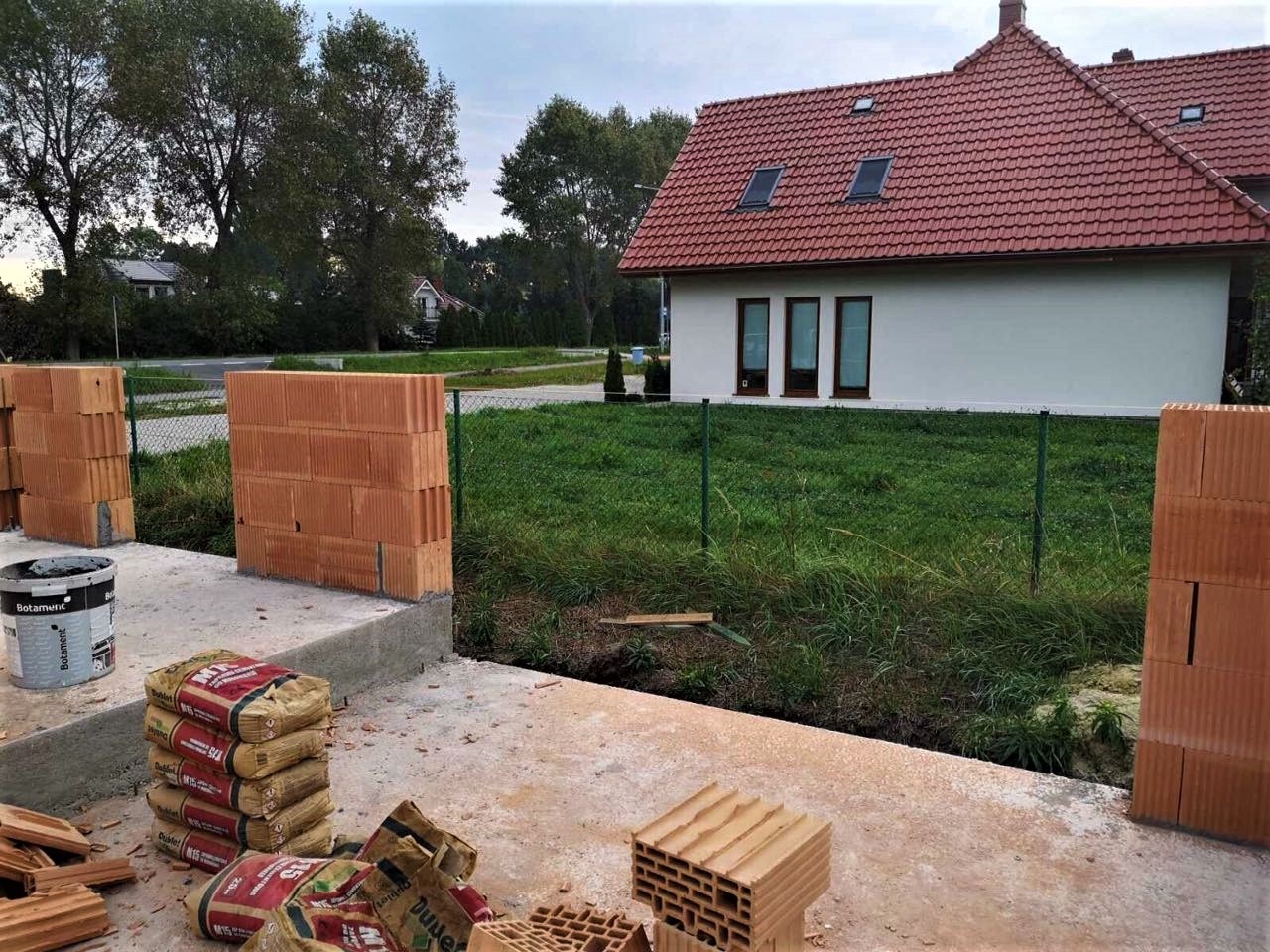 Wznoszenie ścian z czerwonej cegły dziurawki na fundamencie, widoczne worki z zaprawą murarską i wiadro z klejem, w tle dom jednorodzinny z czerwoną dachówką i zielony trawnik.