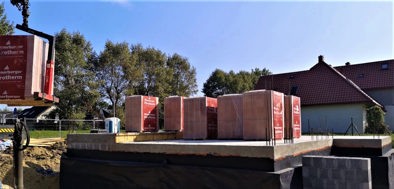 Fundament domu z izolacją pionową i przygotowanym podłożem pod ściany, palety z czerwonymi bloczkami Porotherm dostarczane dźwigiem, w tle dom z czerwoną dachówką.