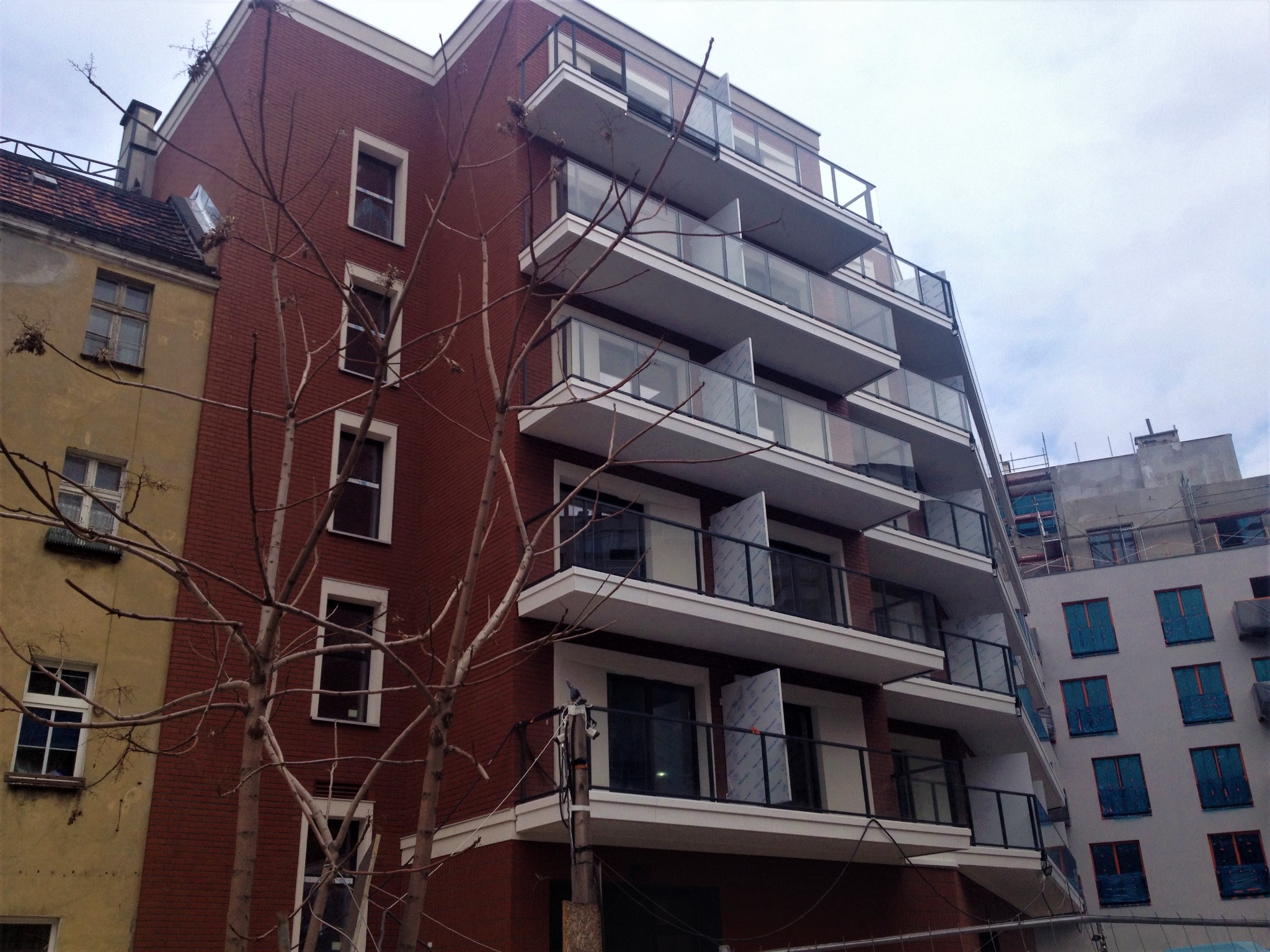 Wielopiętrowy budynek z cegły z balkonami o szklanych balustradach, częściowo w trakcie wykańczania, na tle starszej kamienicy i innego budynku w budowie.