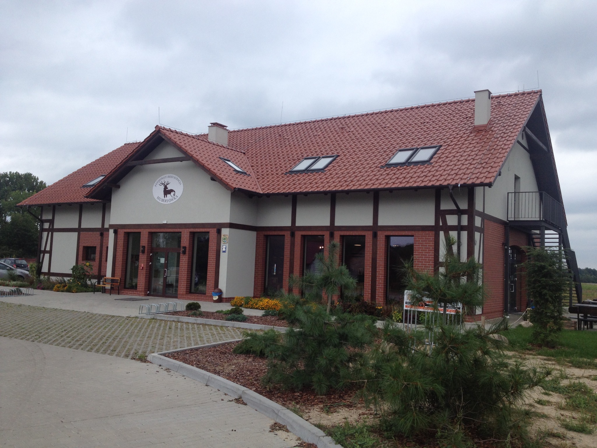 Dwukondygnacyjny budynek z czerwonej cegły i białymi ścianami z elementami drewnianymi, czerwony dach z oknami dachowymi, teren zagospodarowany zielenią, parking z ażurowych płyt betonowych.