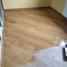 Świeżo polakierowana, błyszcząca podłoga z paneli ułożonych pod kątem, z widoczną kuwetą malarską w prawym dolnym rogu i fragmentem przesuwanych drzwi po lewej stronie.