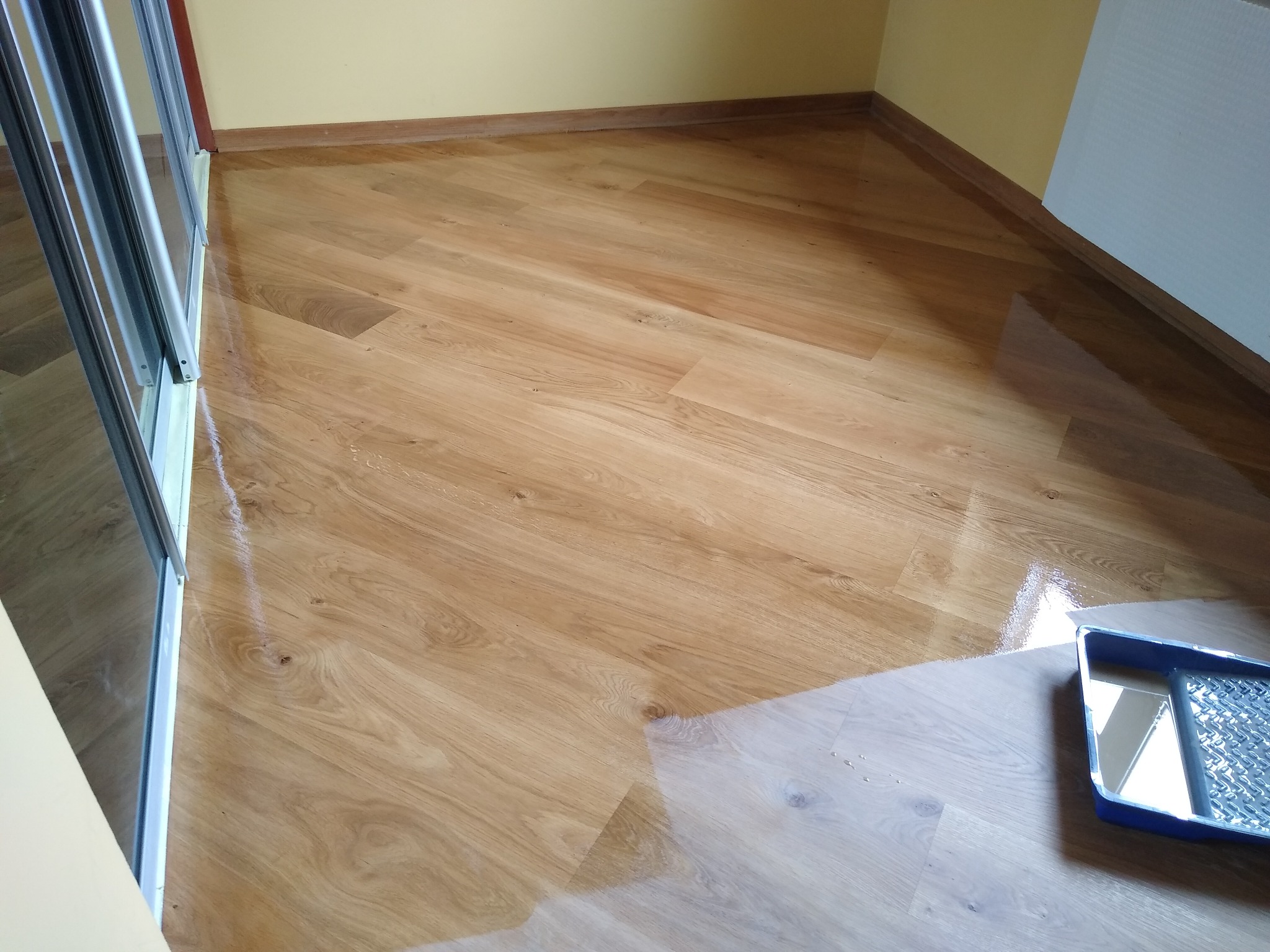 Świeżo polakierowana, błyszcząca podłoga z paneli ułożonych pod kątem, z widoczną kuwetą malarską w prawym dolnym rogu i fragmentem przesuwanych drzwi po lewej stronie.