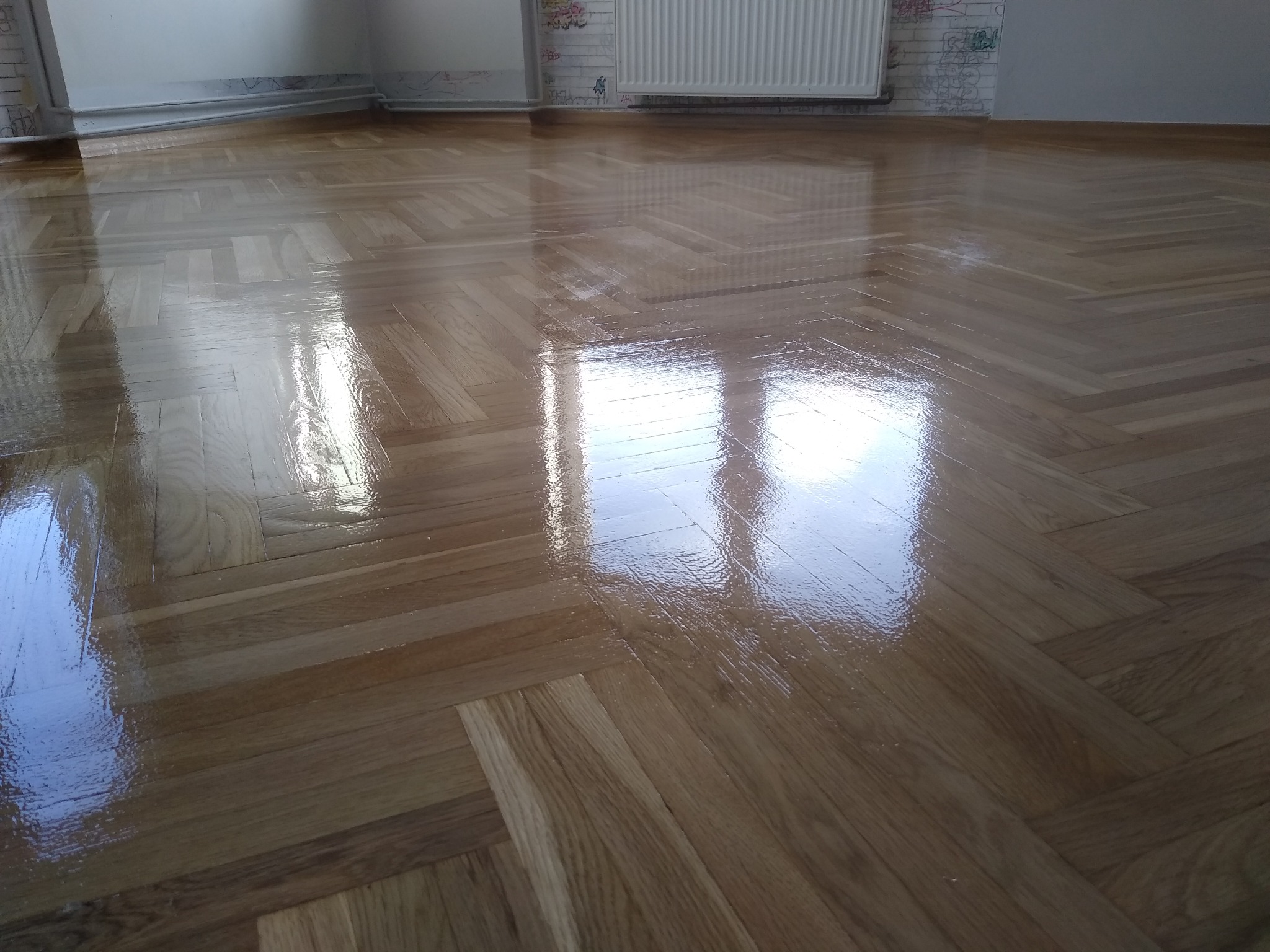 Świeżo polakierowany parkiet ułożony w jodełkę, odbijający światło z okna, z widocznym grzejnikiem i fragmentem ściany z graffiti w tle.