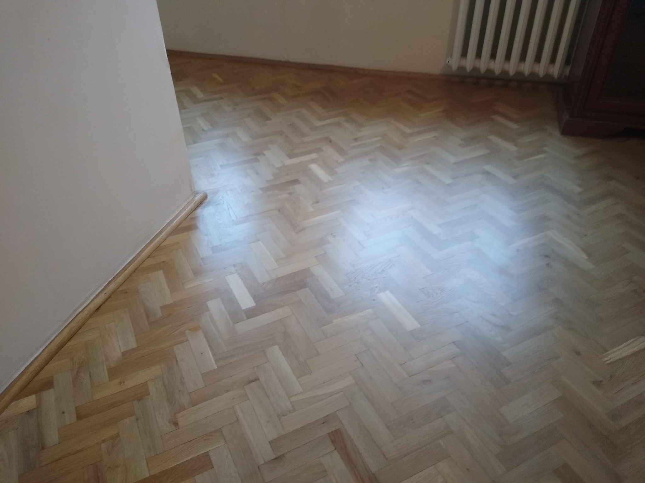Parkiet ułożony w jodełkę, widoczne ślady po cyklinowaniu, przy ścianie listwa przypodłogowa, w tle grzejnik i fragment mebla.