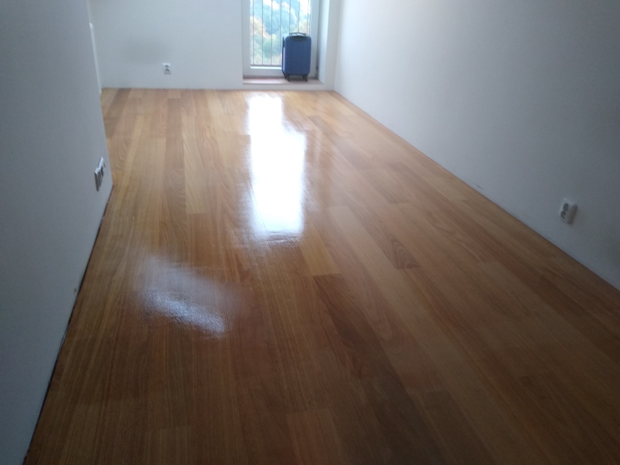 Błyszcząca, świeżo wycyklinowana podłoga drewniana w pustym pokoju z bagażem przy oknie.