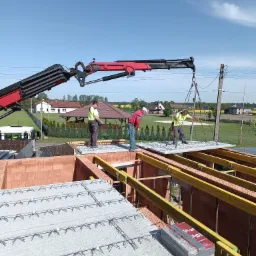 Montaż stropu Teriva z użyciem dźwigu na budowie domu jednorodzinnego z czerwonej cegły, widoczni pracownicy w kamizelkach odblaskowych, błękitne niebo w tle.