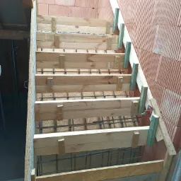 Drewniany szalunek schodów zbrojonych, widoczne pręty zbrojeniowe, ściana z cegły ceramicznej dziurawki w tle.