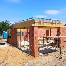 Wznoszenie ścian parterowego domu z czerwonej cegły, widoczne żółte stemple podtrzymujące strop zbrojony stalą, na zewnątrz niebieska toaleta przenośna, teren budowy przygotowany do dalszych prac.