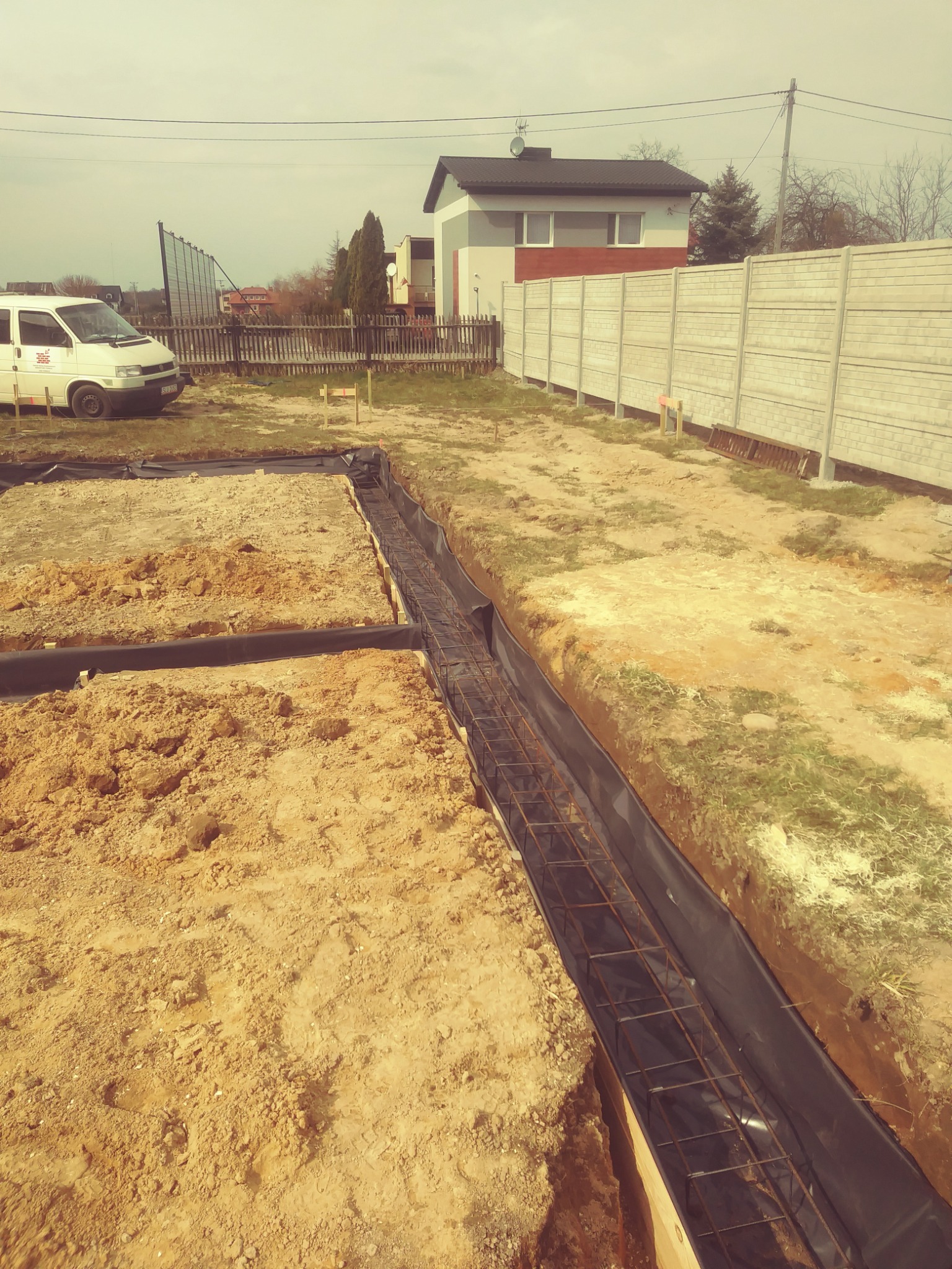 Przygotowane podłoże pod fundamenty domu jednorodzinnego: wykop z szalunkiem i zbrojeniem, zabezpieczony folią, na działce z widocznym samochodem dostawczym i ogrodzeniem.