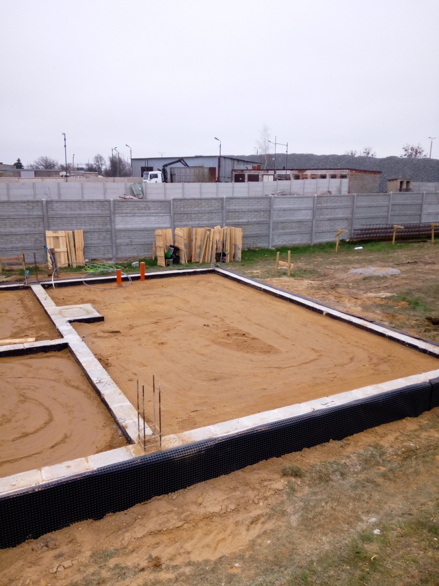 Przygotowany teren pod fundamenty domu z widocznymi zbrojeniami, izolacją pionową i wyprowadzonymi rurami kanalizacyjnymi na tle betonowego ogrodzenia i magazynu.