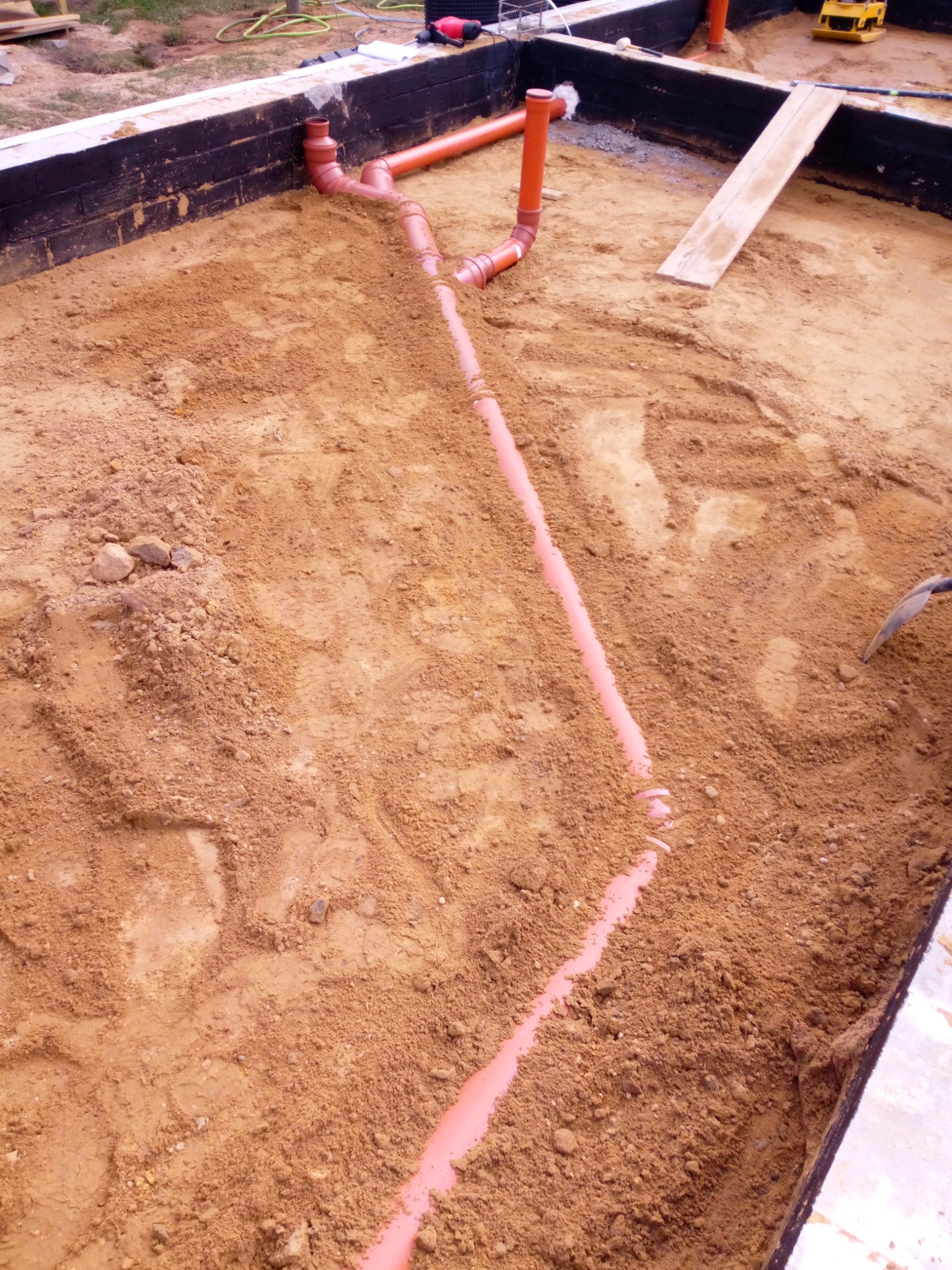 Fundament z ułożonymi rurami kanalizacyjnymi w piasku, widoczne czarne ściany fundamentu oraz częściowo zagęszczarka i deska.