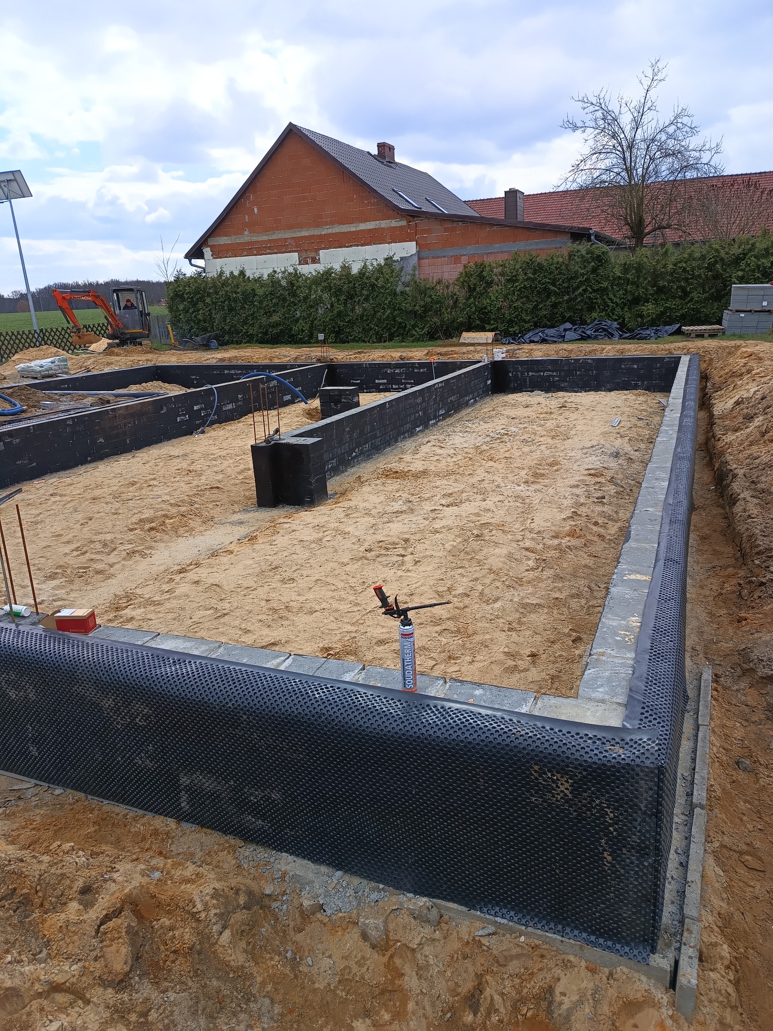 Wykop pod fundamenty domu, z widoczną izolacją przeciwwilgociową, zasypany piaskiem, w tle koparka i budynek w trakcie budowy.